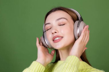 Kusursuz makyajlı, kahverengi dudaklı yeşil arka planda sıradan kazaklı, kablosuz kulaklıklı, kahverengi dudaklı genç bir kadın sesi sever.