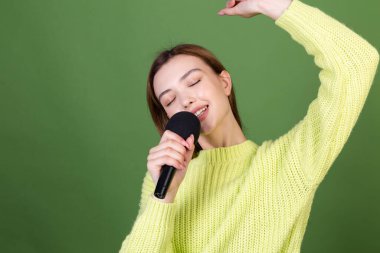 Mükemmel makyajlı, kahverengi dudaklı yeşil arka planda sıradan bir süveter giyen genç kadın mikrofon kapalı gözlerle şarkı söylüyor.