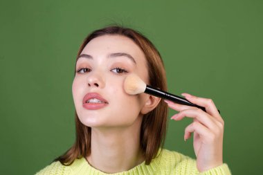 Kusursuz makyajlı, kahverengi dudaklı yeşil arka planda sıradan bir süveter giyen, elinde allık fırçası olan genç bir kadın.