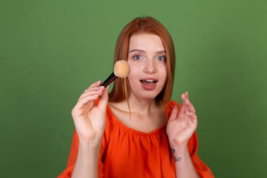 Yeşil arka planda günlük turuncu bluzlu genç kızıl saçlı kadın. Elinde allık fırçası tutan mükemmel, doğal, pürüzsüz bir ten.