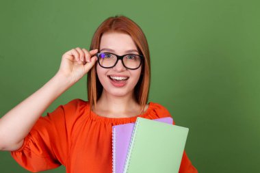 Kırmızı saçlı, sıradan turuncu bluzlu, yeşil arka planda gözlüklü, elinde defterler olan mutlu, neşeli, neşeli bir kadın.