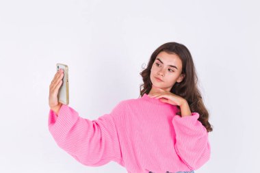 Çilli, hafif makyajlı, beyaz arka planda, cep telefonlu genç ve güzel bir kadın fotoğraf çekiniyor.