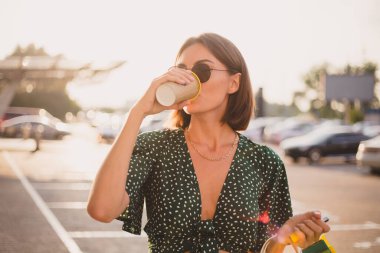 Woman at sunset with colorful shopping bags and coffee at parking lot  by shopping mall