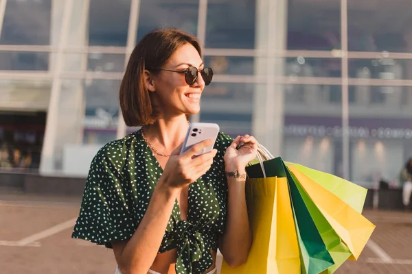 Woman at sunset with colorful shopping bags and parking lot by shopping mall happy with mobile phone