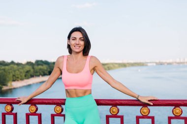 Günbatımında modern briç sahasında spor elbisesi giyen güzel bir kadın. Nehir manzaralı mutlu bir gülümseme.