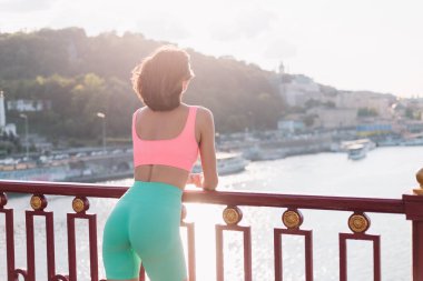 Günbatımında nehir manzaralı modern köprüde spor elbisesi giyen sporcu kadın yaz havasının tadını çıkarıyor.