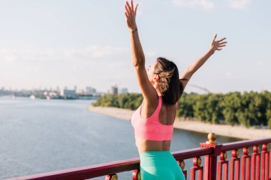 Günbatımında modern köprüde spor elbiseleri giyen sportif bir kadın nehir manzaralı el kaldırıyor yaz havasının tadını çıkarıyor, özgürlüğüne güveniyor.