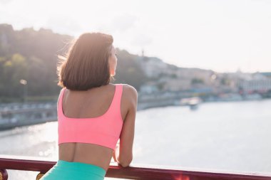 Günbatımında nehir manzaralı modern köprüde spor elbisesi giyen sporcu kadın yaz havasının tadını çıkarıyor.