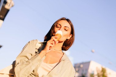 Yaz günbatımında kadın dışarıda dondurma yiyor. Şehir caddesinde neşeli, neşeli bir dondurma yiyor.