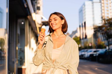 Yaz günbatımında kadın dışarıda dondurma yiyor. Şehir caddesinde neşeli, neşeli bir dondurma yiyor.