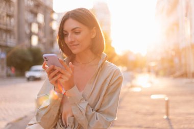 Yazın gün batımında kadın dışarıda cep telefonuyla mutlu bir gülümsemeyle ekranlara bakın.