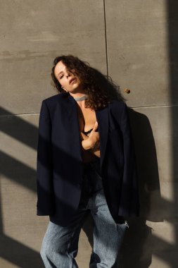 Stüdyoda ıslak saçlı güzel bir kadın, siyah büyük beden ceket ve parlak bir kolye giyiyor, doğal güneş ışığı, duvarda poz veriyor.