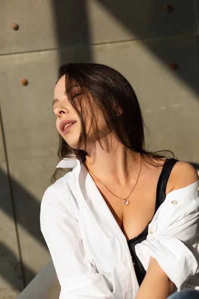 大きな胸を持つかなり若い女性は スタジオで白い椅子にポーズ 後ろの鋼壁 ジーンズと白いシャツを着て 自然な暖かい日没の光 — ストック写真