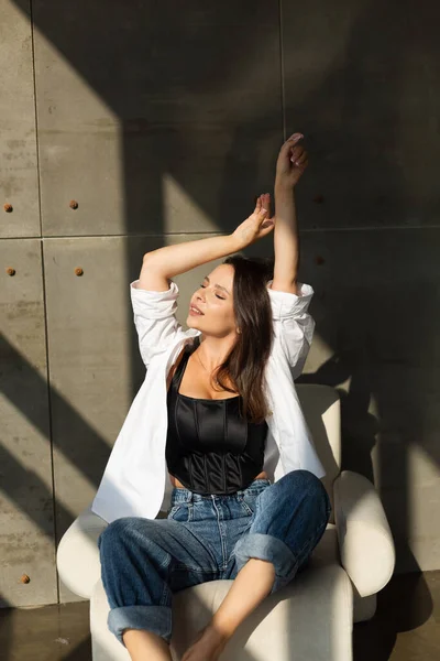 Pretty young woman with big breast posing on white chair in studio, steel wall behind, wearing jeans and white shirt, natural warm sunset light