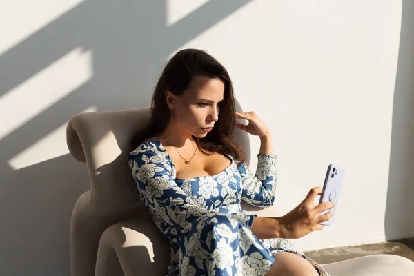 美しいです若いです女性で大きな胸ポージングでスタジオ 長い夏のフィッティングドレスを着て 自然な暖かい日没の光 椅子に座って写真を撮る自己撮影電話で — ストック写真
