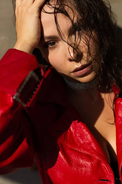 Pretty young woman with wet hair posing in studio, wearing leather red cloak and shiny necklace, natural warm sunset light, standing by wall