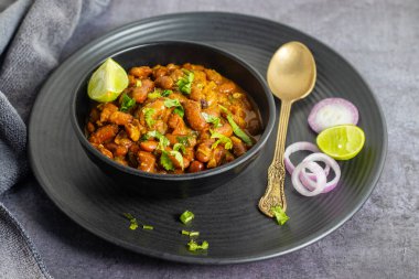 Limonlu soğan ve kaşıkla servis edilen siyah kâsede kişniş ile süslenmiş baharatlı rajma körisinin üst görüntüsü..