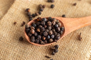 Tahta kaşık siyah biberlerle dolu, beyaz ahşap bir masa yüzeyine saçılmış, rustik çuval bezi kumaş üzerinde dinleniyor..