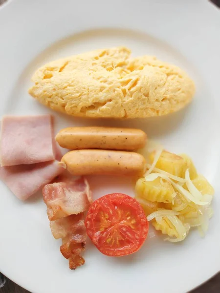 Beyaz tabakta kahvaltıda çırpılmış yumurta, sosis, jambon, domates ve yakınlaştırma var..
