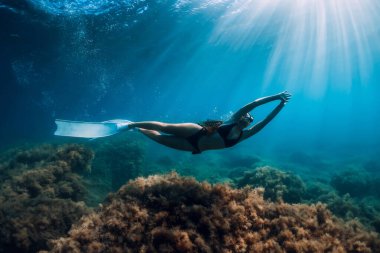 Beyaz yüzgeçli özgür bir kadın güneş ışınlarıyla deniz yosunlarının üzerinden süzülüyor. Okyanusta serbest dalış