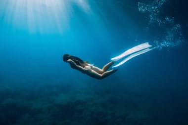 Özgür yüzgeçli genç bir kadın güneş ışınlarıyla denizin altında süzülüyor. Şeffaf okyanusta serbest dalış