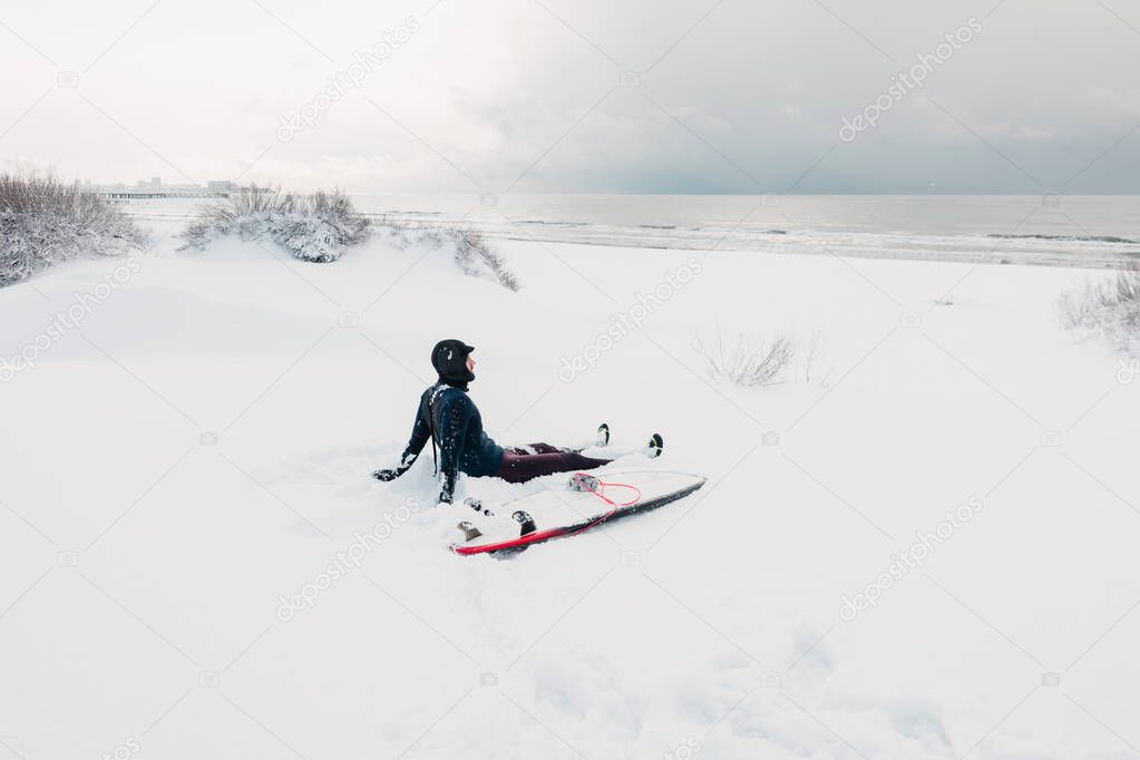 Cold winter and surfer sitting on snow beach with surfboard. Winter day with surfer in wetsuit.