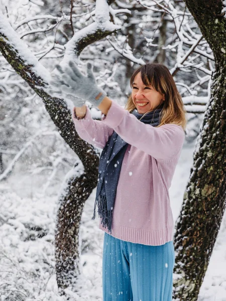 Happy young woman standing among snowy trees in winter forest and enjoying first snow. Smiling woman play with snow
