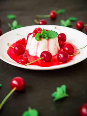 Koyu arkaplanda beyaz tabakta rakun sütü, kiraz şurubu ve böğürtlenli panna cotta, vejetaryen tatlı.