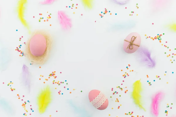 Ester frame composition with colorful eggs, feathers and confetti on white background. Flat lay, top view