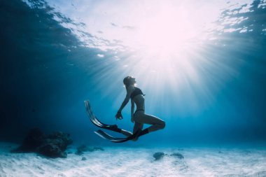 Genç kadın yüzgeçleriyle kumlu denizde süzülüyor. Mavi okyanusta ve güneş ışığında serbest dalış