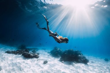 Genç kadın yüzgeçleriyle kumlu denizde süzülüyor. Mavi okyanusta ve güneş ışığında serbest dalış