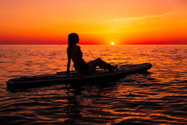 明るい夕日や日の出と静かな海でパドルボードに立つ女の子 女性は海でボード上でリラックス — ストック写真