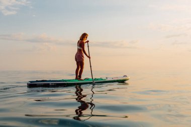 25 Haziran 2021. Anapa, Rusya. Akşamları sakin denizde sportif kızlar ayakta yüzer. Kadınlar Red Paddle SUP sörf tahtasında.