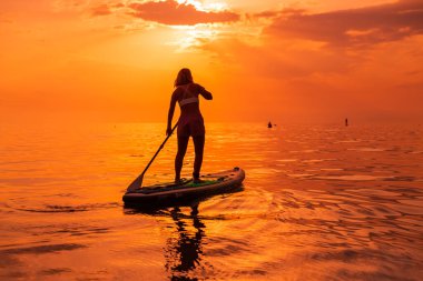 25 Haziran 2021. Anapa, Rusya. Sportif kadın günbatımında ya da gündoğumunda sakin denizde ayakta kürek çekiyor. Kırmızı yüzgeçli kadın yemek masasında ve günbatımında yansımalı