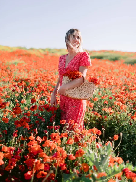 Sensual young woman in a poppy field at sunset. Concept of freedom with soft colors. Woman in red dress and handbag with flowers