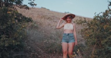 Çekici bir şekilde kameraya gülümseyen bir kadın. Şık bir genç kız poz veriyor ve gün batımını seyrediyor. Ilık ve modaya uygun bir şapka takıyor. Mutlu bir kadının doğal güzelliği.