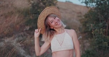 Çekici bir şekilde kameraya gülümseyen bir kadın. Şık bir genç kız poz veriyor ve gün batımını seyrediyor. Ilık ve modaya uygun bir şapka takıyor. Mutlu bir kadının doğal güzelliği.