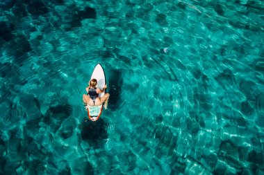 Şeffaf turkuaz okyanusta sörf tahtasında oturan sörfçü kız. Sörfçü kadınla hava manzarası