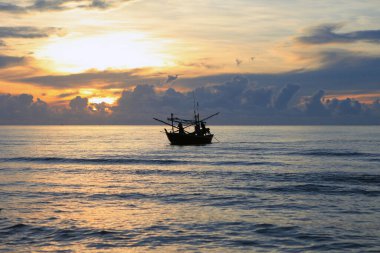 Küçük bir balıkçı teknesinin silueti Denizde balık tutuyor, Sabah güneş yükseliyor ve bulutlar güzel, ılık turuncu renkler. 
