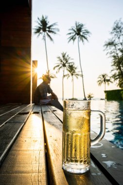 Ahşap bir güverteye buz ve soğuk içeceklerle doldurulmuş bir bardak bira. Arka planda siluet Adam ve hindistan cevizi ağaçlarının güneş ışınları var. Yalnızlık ve üzüntü konsepti.