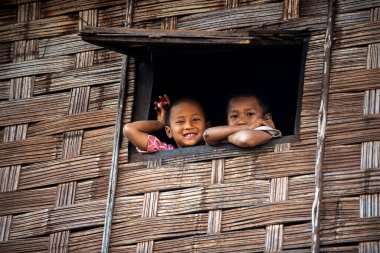 Chiang Mai / Tayland - 16 Ocak 2016: Ahşap bir evin penceresinde duran iki erkek çocuğu. Duvar bambudan yapılmış, çocuk gülümsüyor ve evden dışarı bakıyor. Kuzey Tayland 'daki kırsal köyler