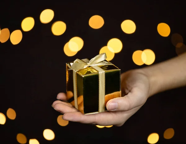 Рука тримає золоту подарункову коробку з золотою стрічкою, іскровим фоном, Різдвом або Днем народження, подарункова концепція з простором для тексту — стокове фото