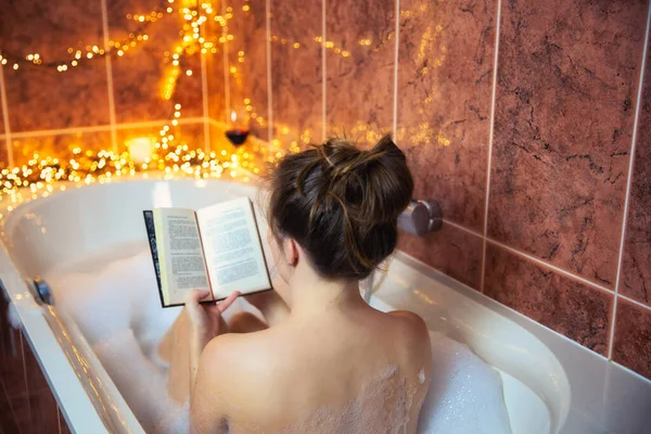 Ung vacker kvinna läser en bok och dricker rött vin i badkaret med bubbelskum, dekorerad med färgglada ljus, avkopplande och spa koncept — Stockfoto