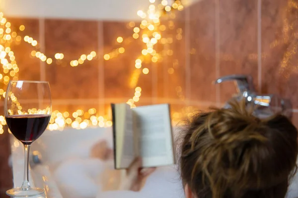 若い美しい女性が本を読んで、泡泡と浴槽で赤ワインを飲みます,カラフルな照明で飾られました,リラックスしてスパのコンセプト — ストック写真