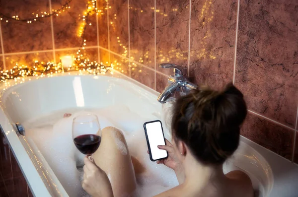 Ung kvinna som håller ett glas rött vin och använder sin Smartphone i badkaret med bubbla skum och ljus på bakgrunden, kopiera utrymme — Stockfoto