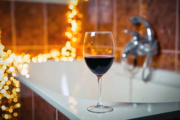 Close up of wine glass with red wine near bathtub with lights on the background, relaxtion and spa concept — Stock Photo, Image