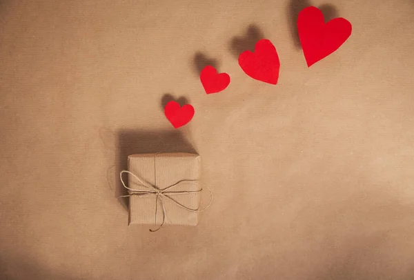 Vysoký úhel pohledu na prostý ekologicky šetrný hnědý papír zabalený pro Valentines Day dárek s různými červenými srdci na hnědém papíře pozadí kopírovat prostor — Stock fotografie