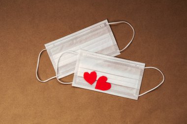 İki kırmızı kalp ve beyaz yüz koruma maskesi, Sevgililer Günü ve Coronavirus konsepti kahverengi kağıttan arka planda. Boşluğu kopyala