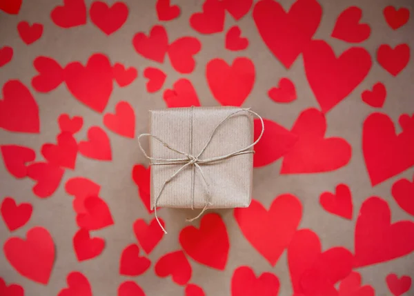 Dárkové balení zabalené v kraft papíru obklopený červenými srdci na hnědém prostém papíře pozadí, Valentines Day koncept top view moderní design, kopírovací prostor — Stock fotografie