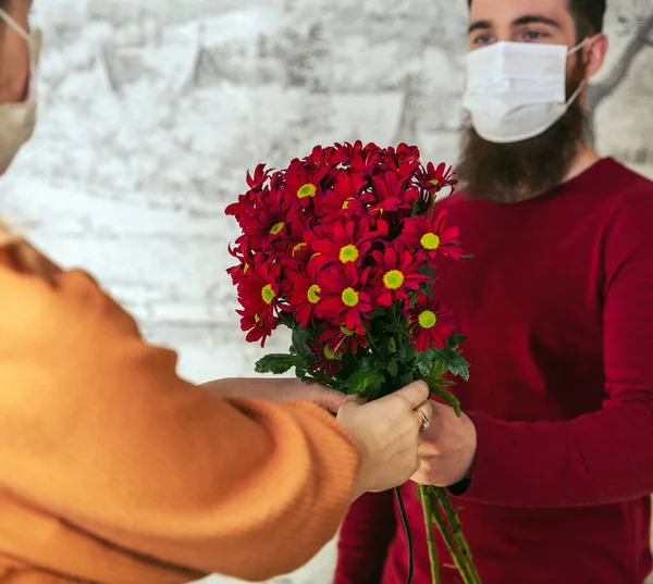 Молодий романтичний люблячий чоловік, одягнений в захисну маску для коронавірусу і дає молодій жінці букет квітів, Ковід-19, День святого Валентина, день народження. — стокове фото